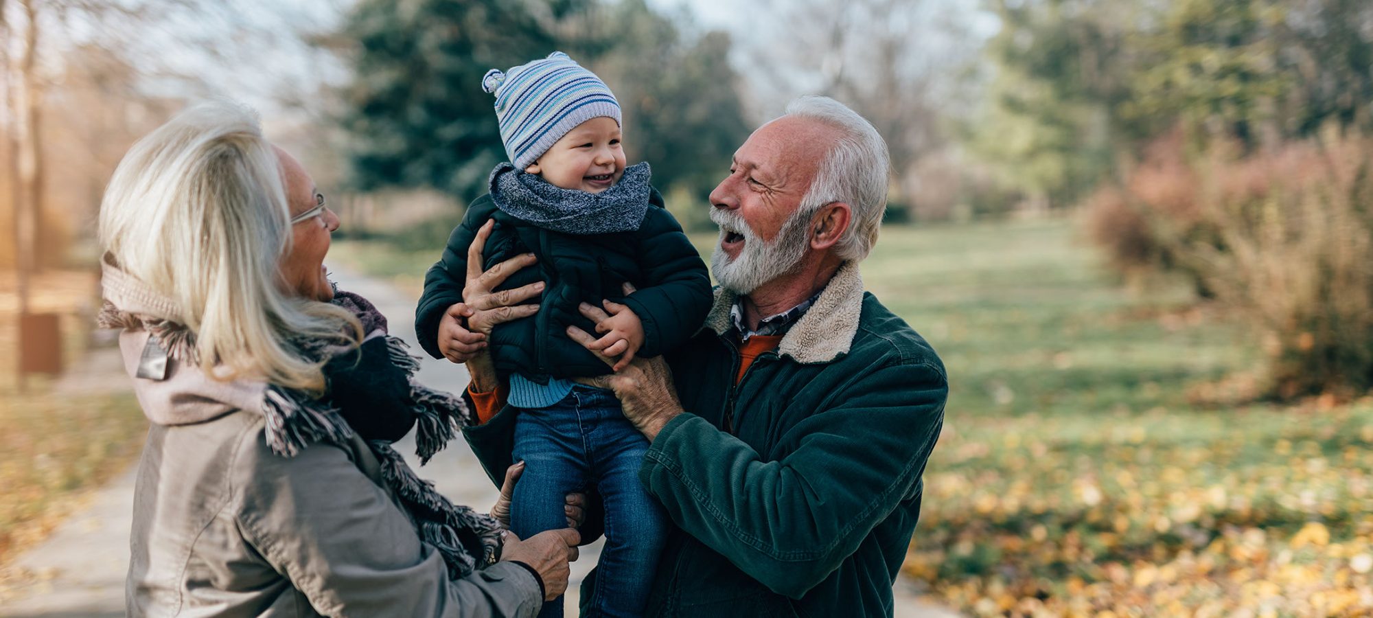 Lykkelig, pent eldre par, ektemann og kone, går og leker med sitt bedårende barnebarn i offentlig bypark.